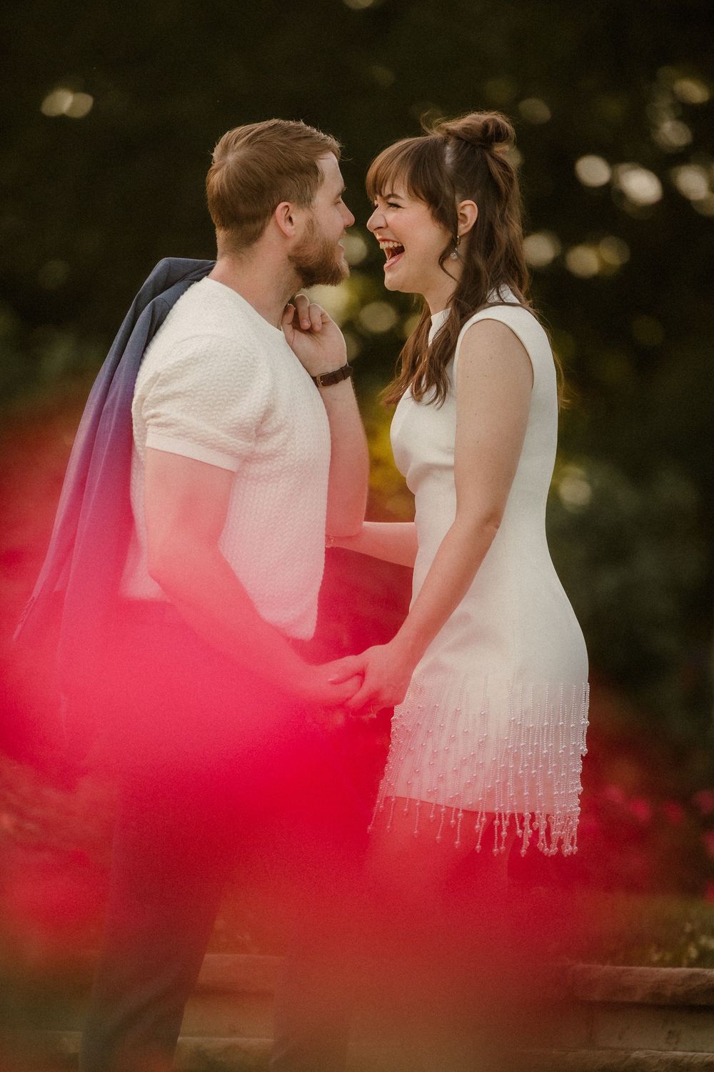 A joyful couple laughing together outdoors with a red-tinted foreground element creating a romantic atmosphere. Overland Park Arboretum.