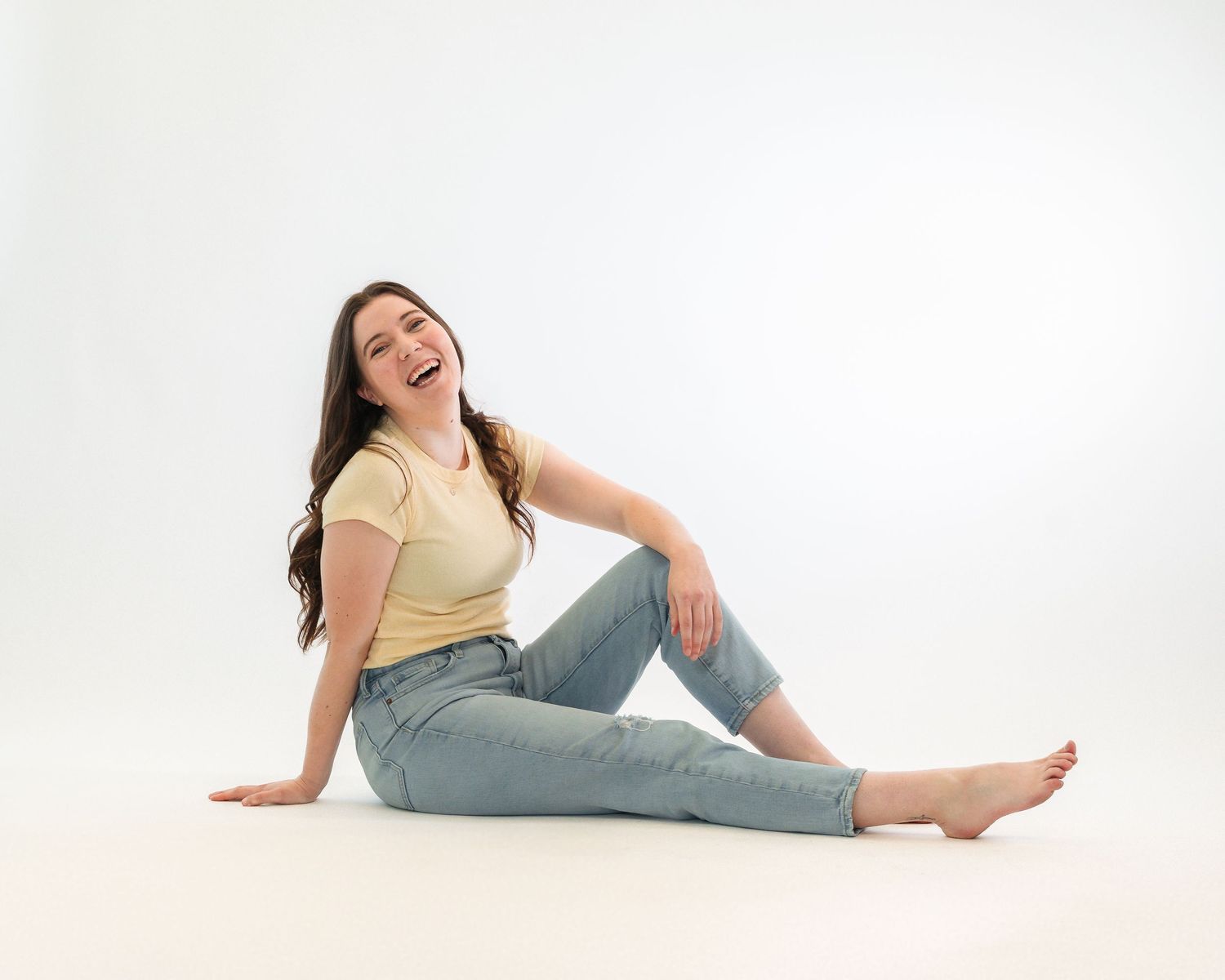 A person wearing a yellow shirt and jeans sits casually on a white studio floor.