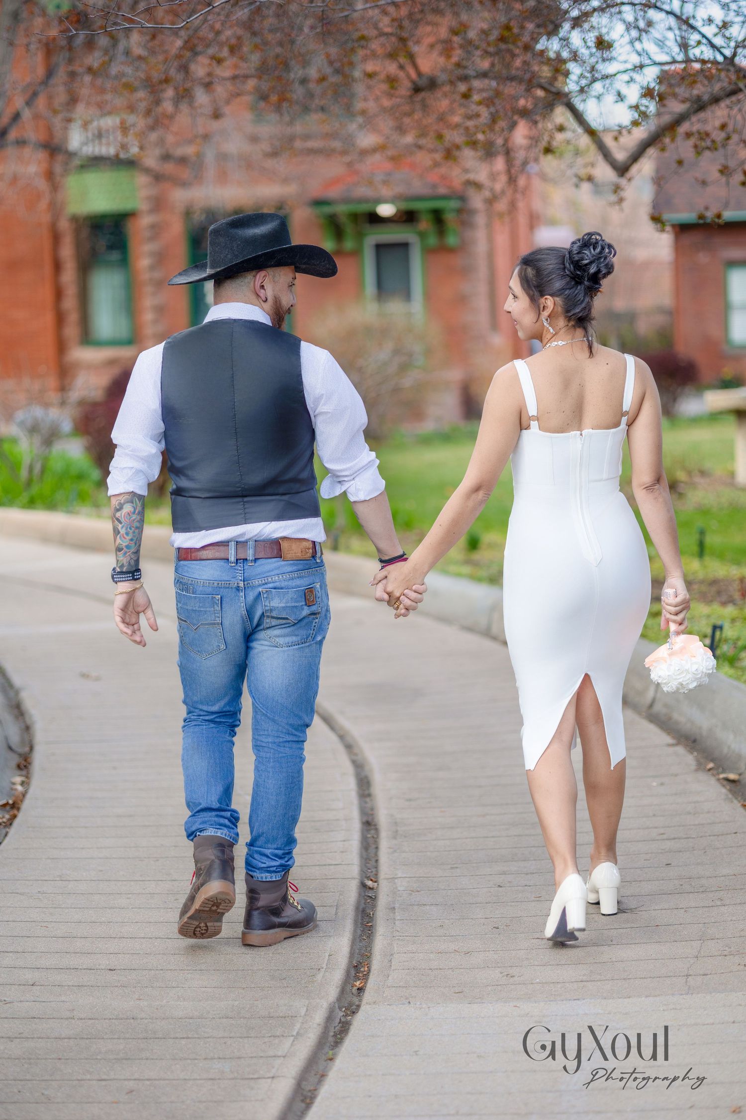 A couple walking hand in hand on a brick pathway, with the person in a hat and vest leading the way.