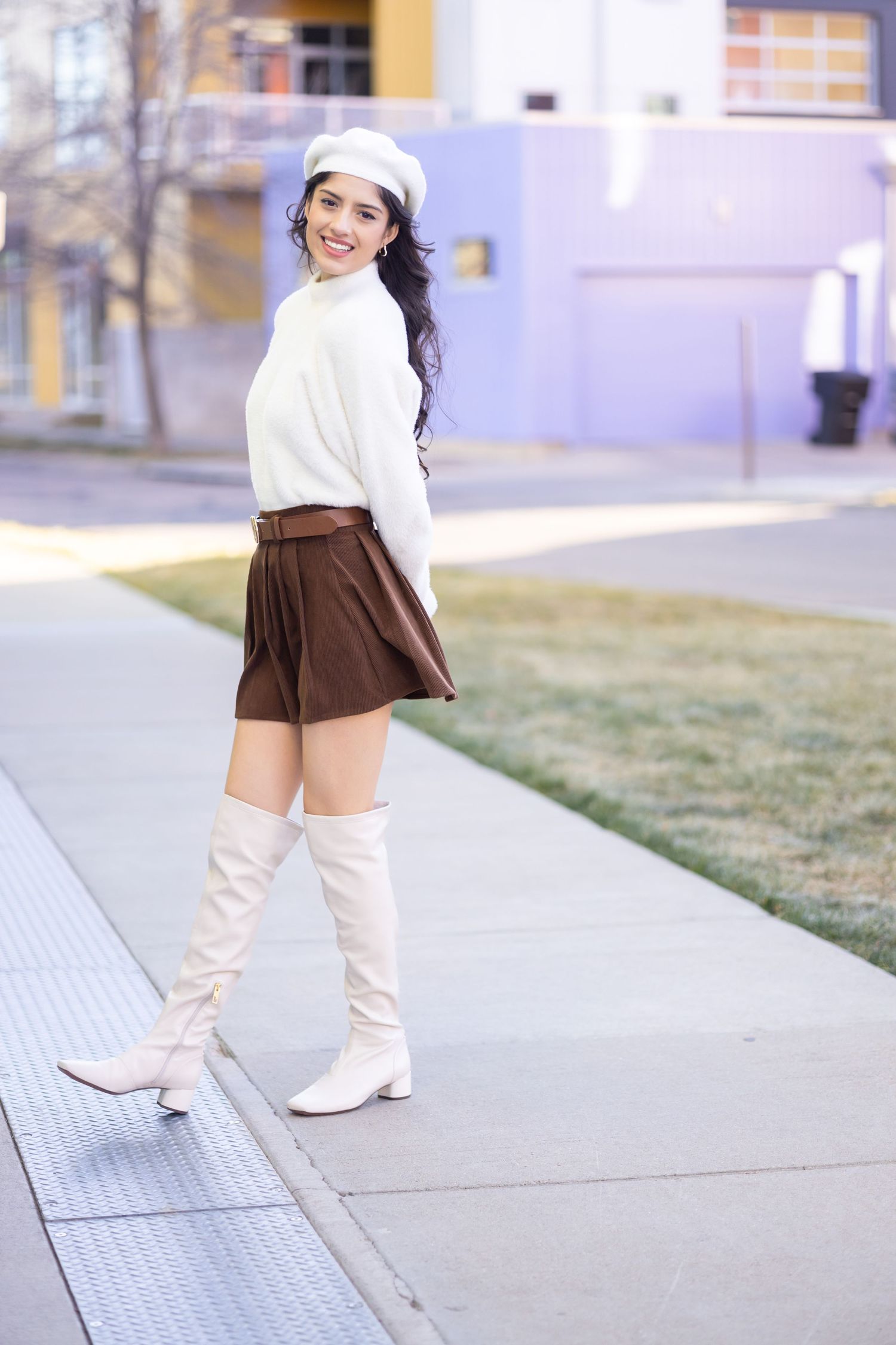 Stylish winter outfit featuring white sweater, brown skort, cream knee-high boots and matching beret on city sidewalk.