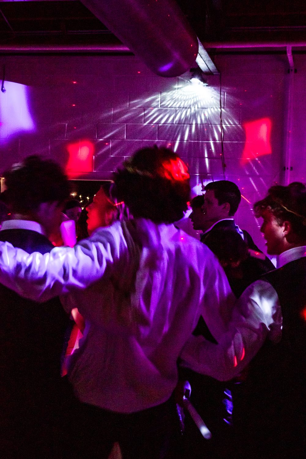 People dance and celebrate at a nightclub illuminated with purple and red lights while strobes create geometric patterns.