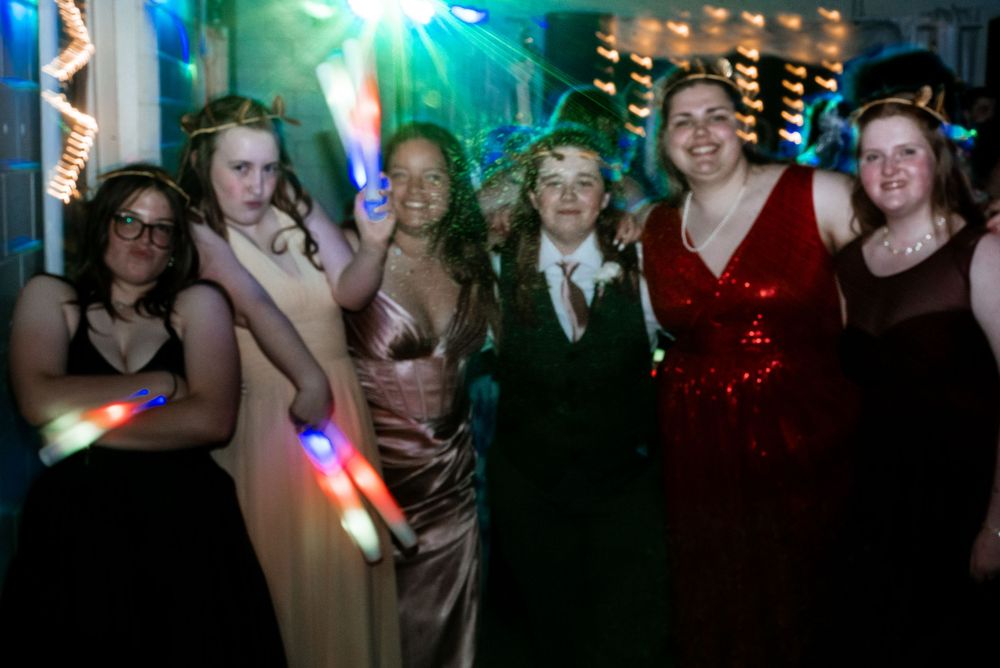 A lively group celebrates at a nighttime party or dance with colorful lights and formal attire in the background.