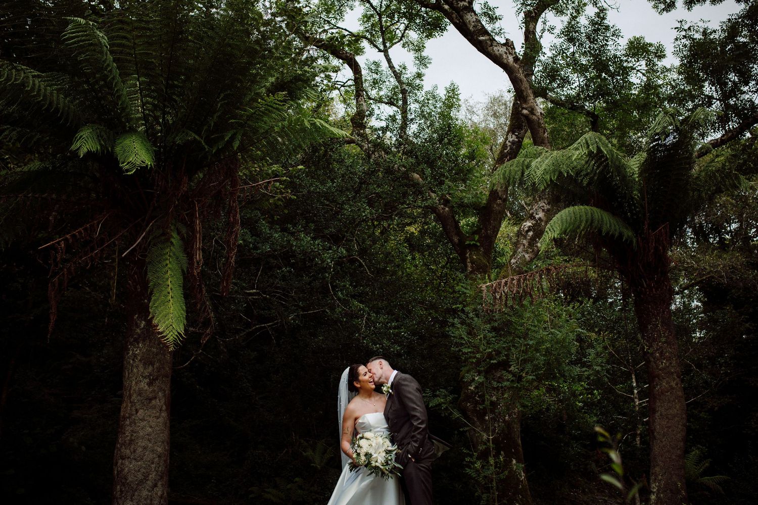 Two people embrace in a romantic moment surrounded by lush palm trees and greenery in a tropical garden setting.