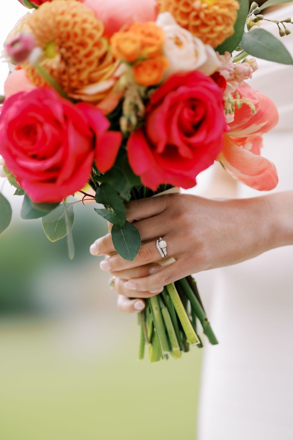 A vibrant bridal bouquet featuring red roses, orange dahlias, and greenery held by hands with an elegant wedding ring at Hall of Springs.