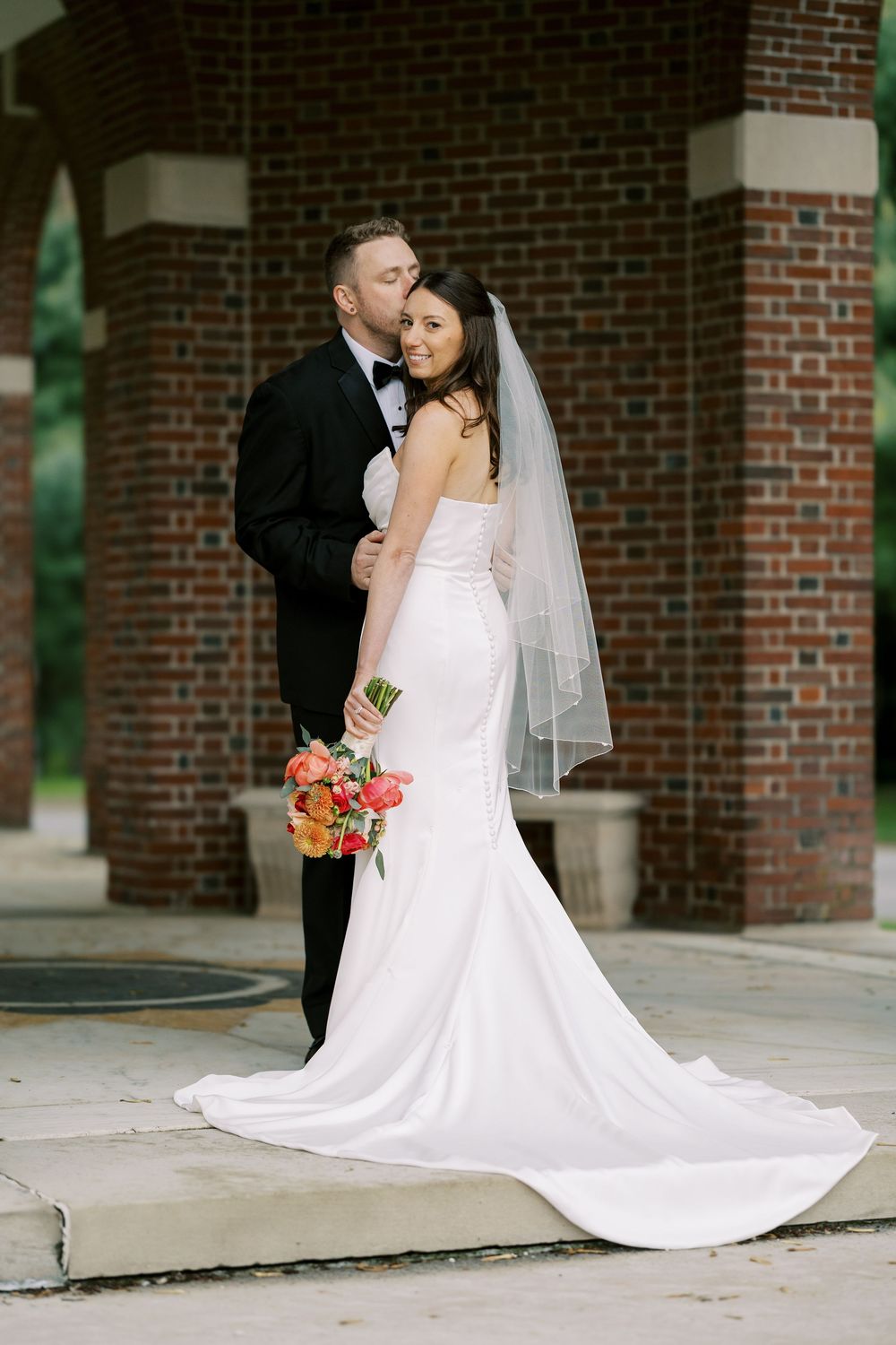 A loving couple in wedding attire embrace outside a brick building, with the bride holding a coral bouquet at Hall of Springs.