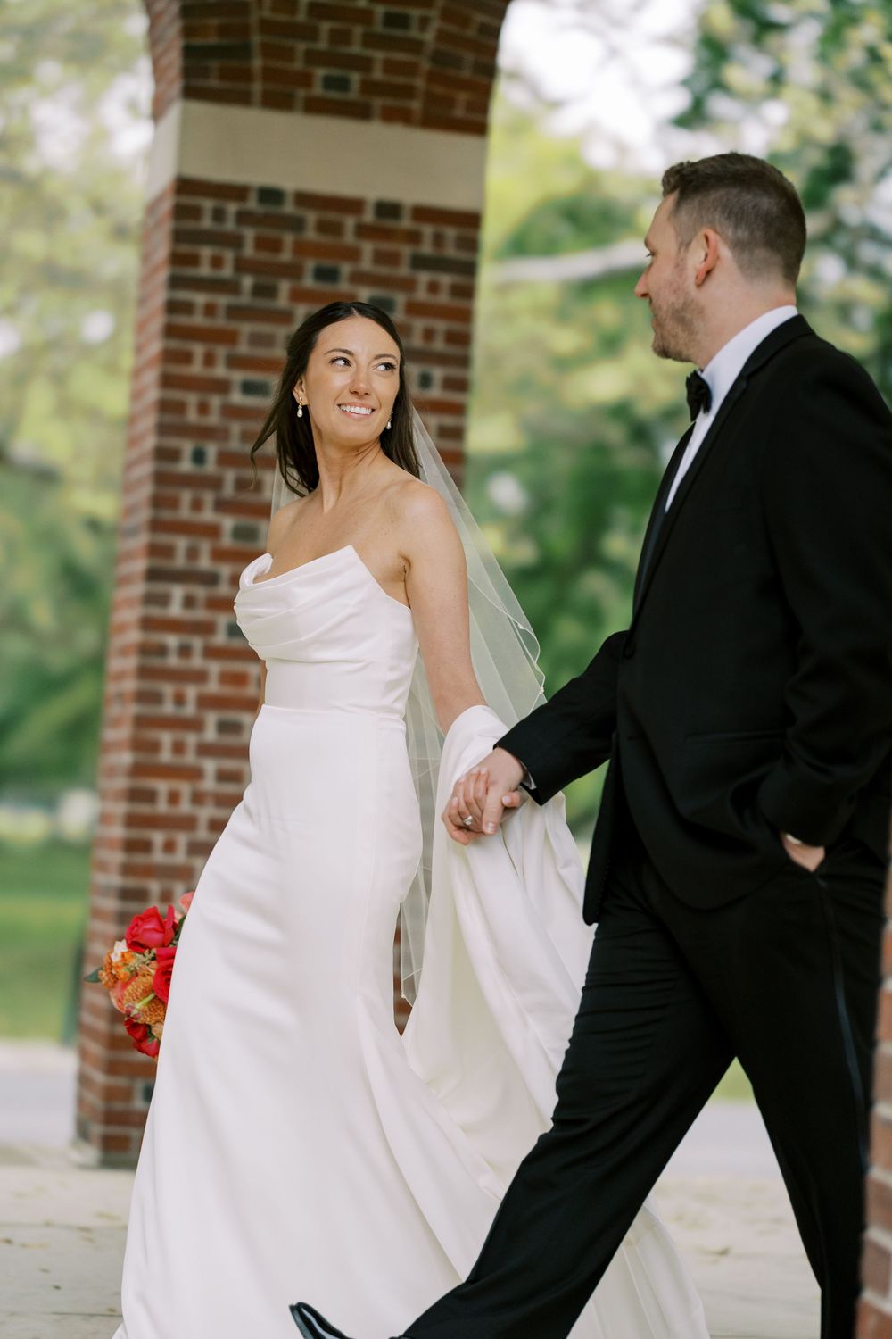 A bride in a strapless white gown and her groom in a black tuxedo share a playful moment near a brick pillar outdoors at Hall of Springs.