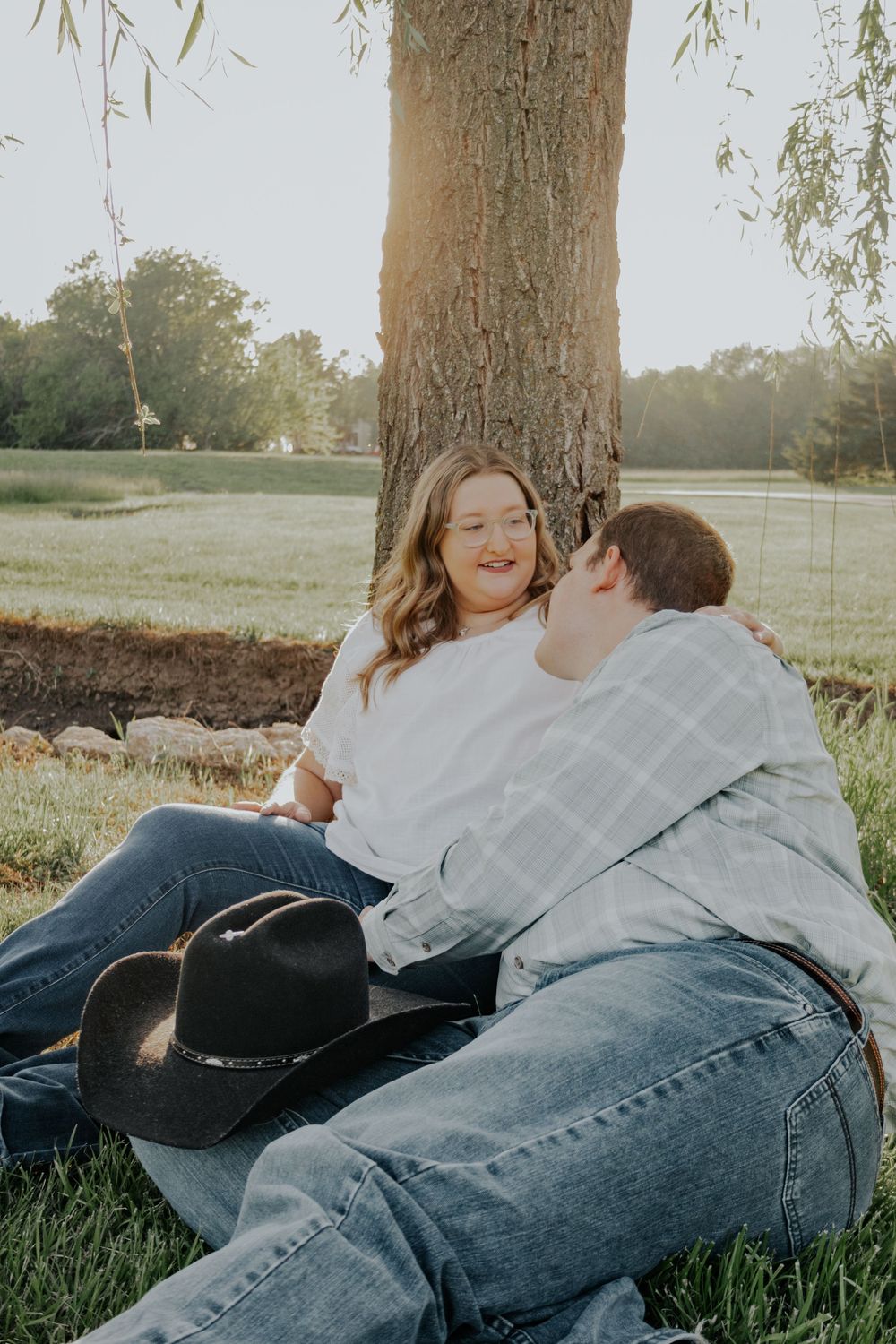 A couple sitting together under a large tree in a grassy field at sunset with a cowboy hat beside them.