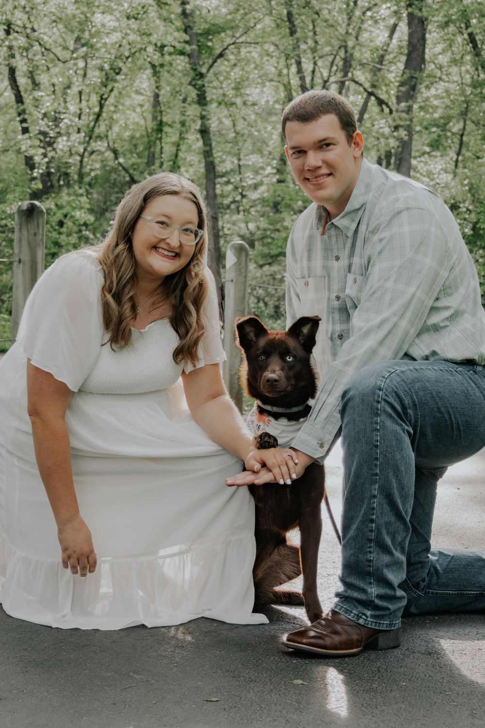 A couple poses for their engagement photo in a wooded setting with a black dog between them.