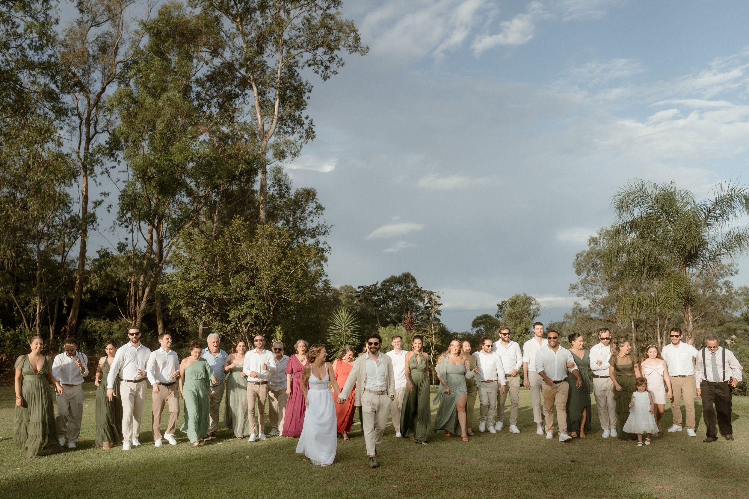 Large wedding party group photo on a lawn with tall trees and cloudy sky backdrop.