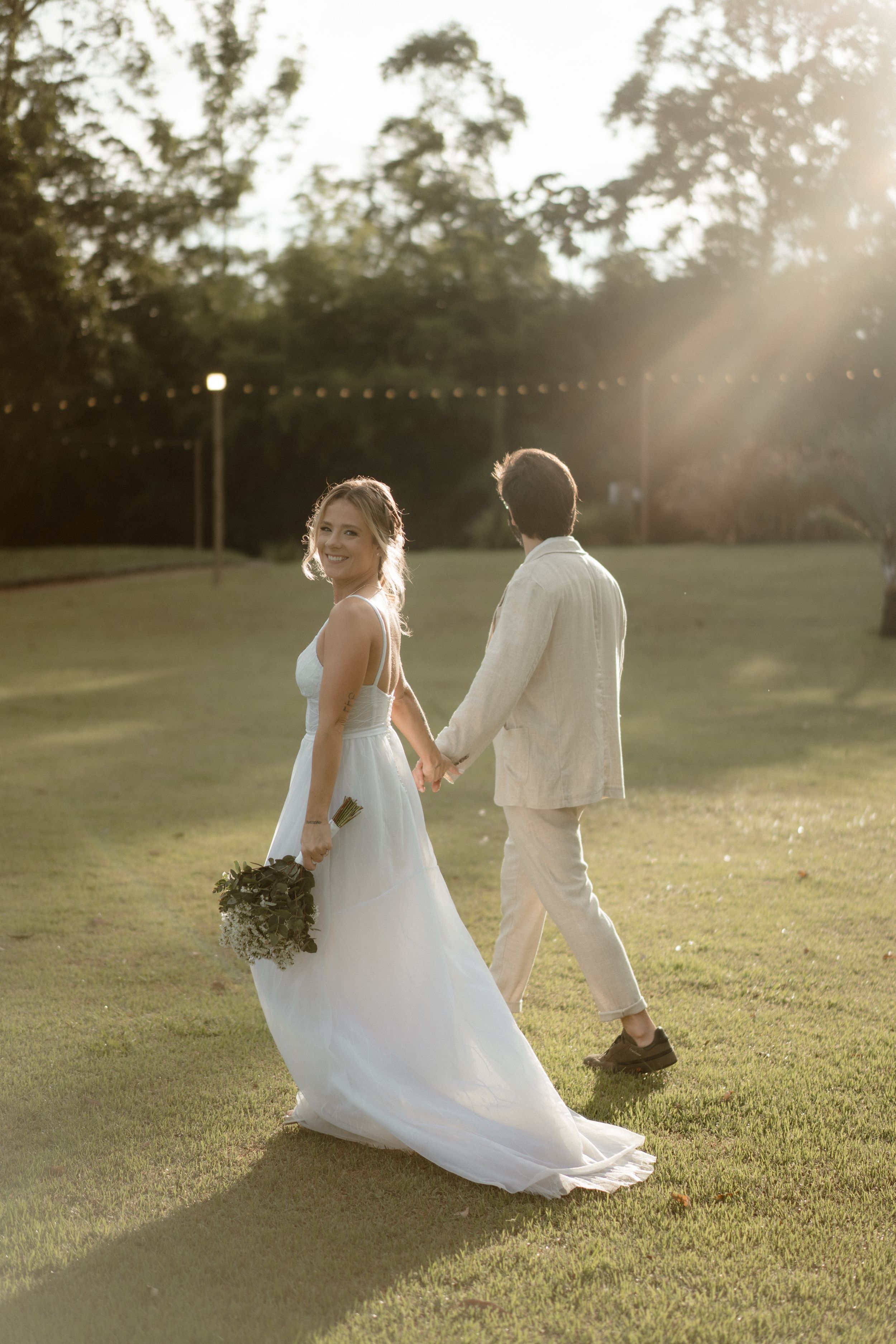 Sunset wedding portrait of couple walking hand in hand on grass with flowing white dress.