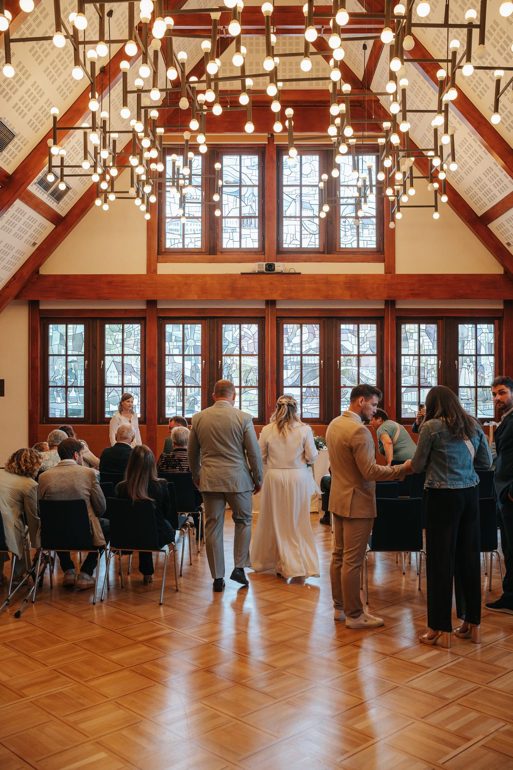 Guests gather in an elegant venue with high ceilings, wooden beams, and hanging string lights during an indoor celebration.