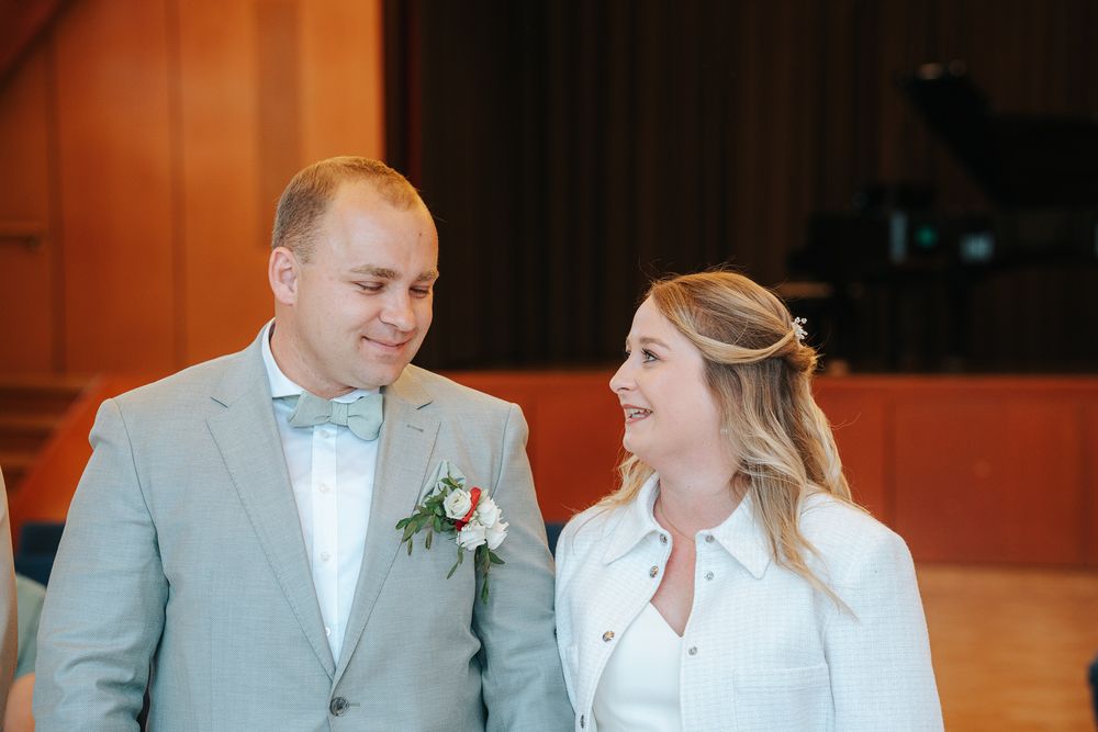 Happy couple at indoor wedding venue wearing light grey suit and white dress with turquoise bow tie.