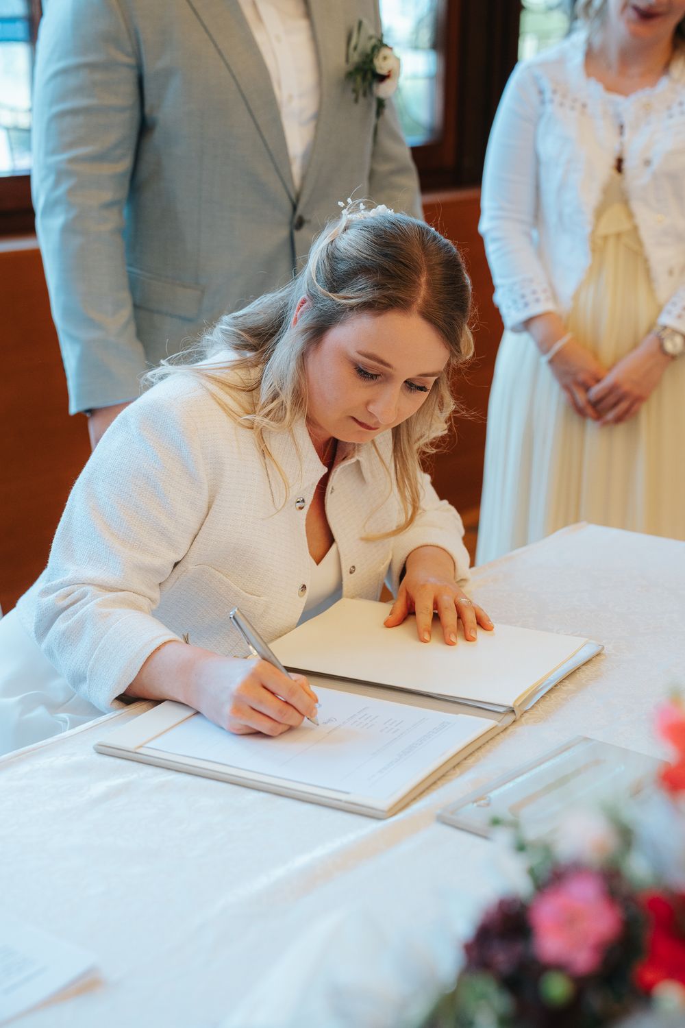 Person signs wedding registry book at ceremony wearing white dress while others look on.