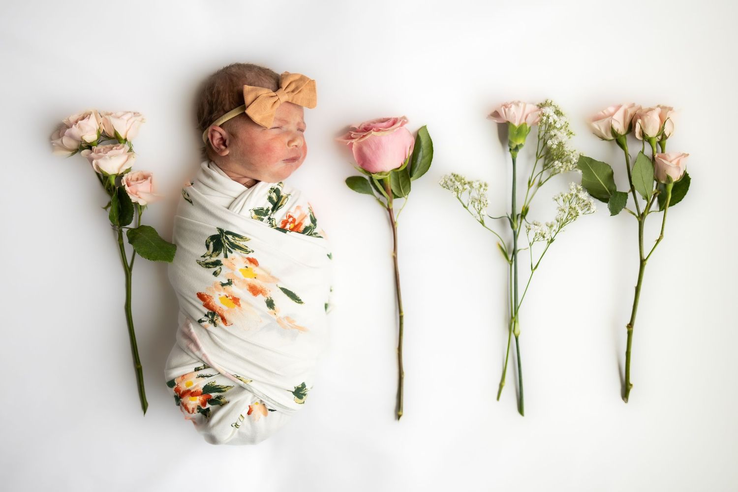 Newborn baby wrapped in floral swaddle blanket lying beside pink roses and white carnation flowers on white background.