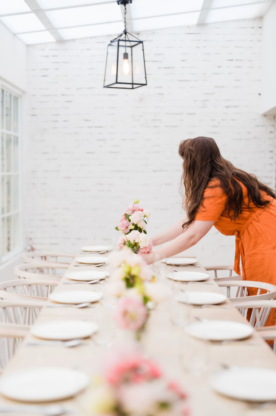 An orange-dressed person sets a long rustic table with white plates and floral arrangements in a bright white dining space at Sugar Pea Cafe