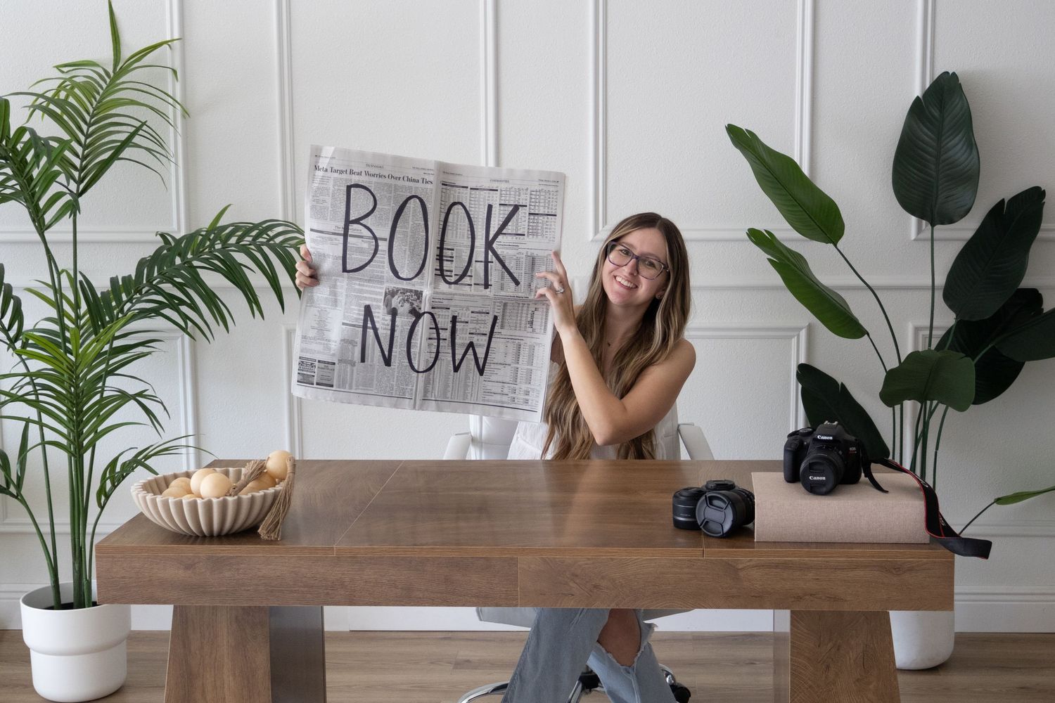 Photographer branding image at a desk with plants, camera equipment, and a newspaper reading 'book now.'