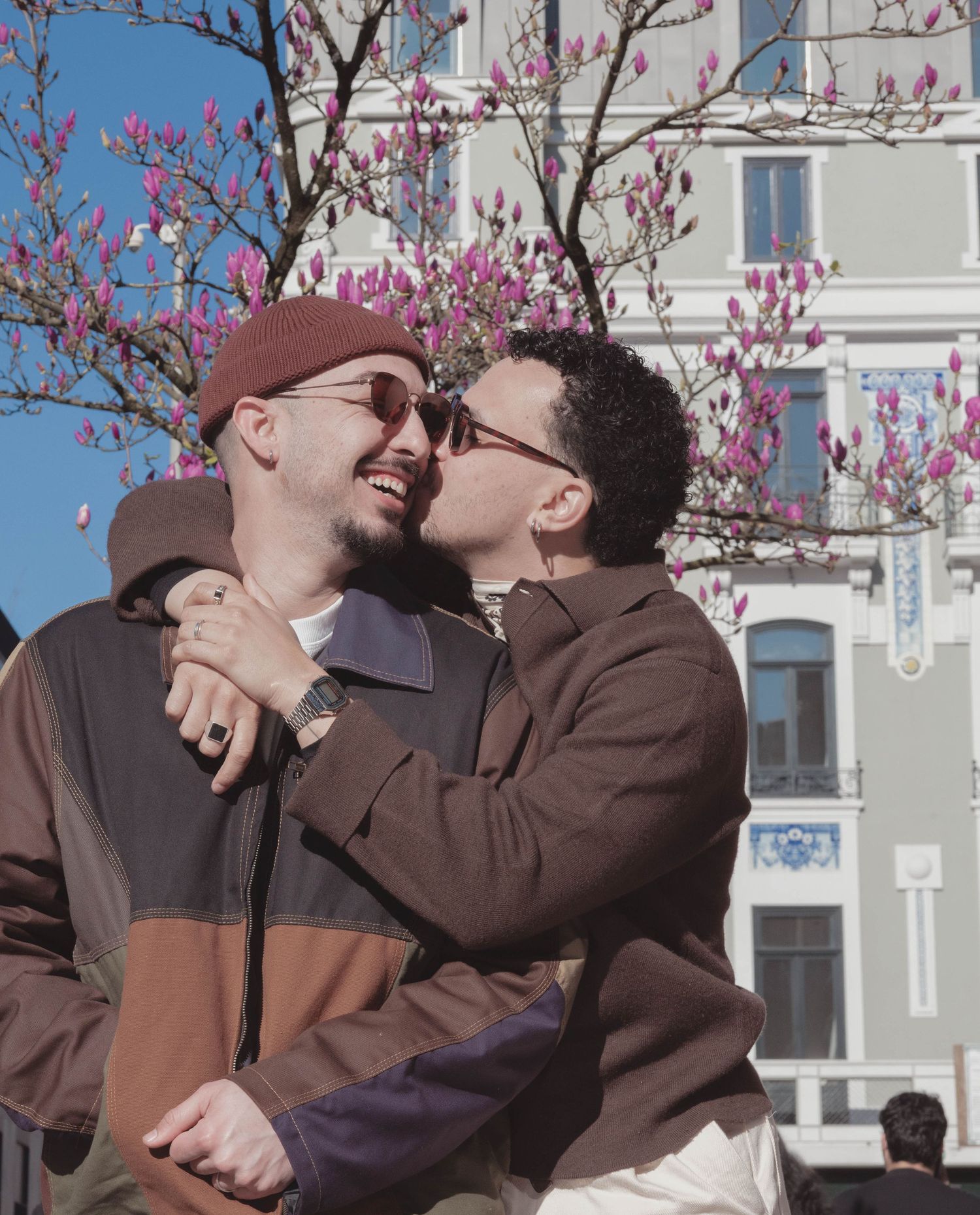 Two people sharing an affectionate moment under blooming cherry blossoms against a white building.