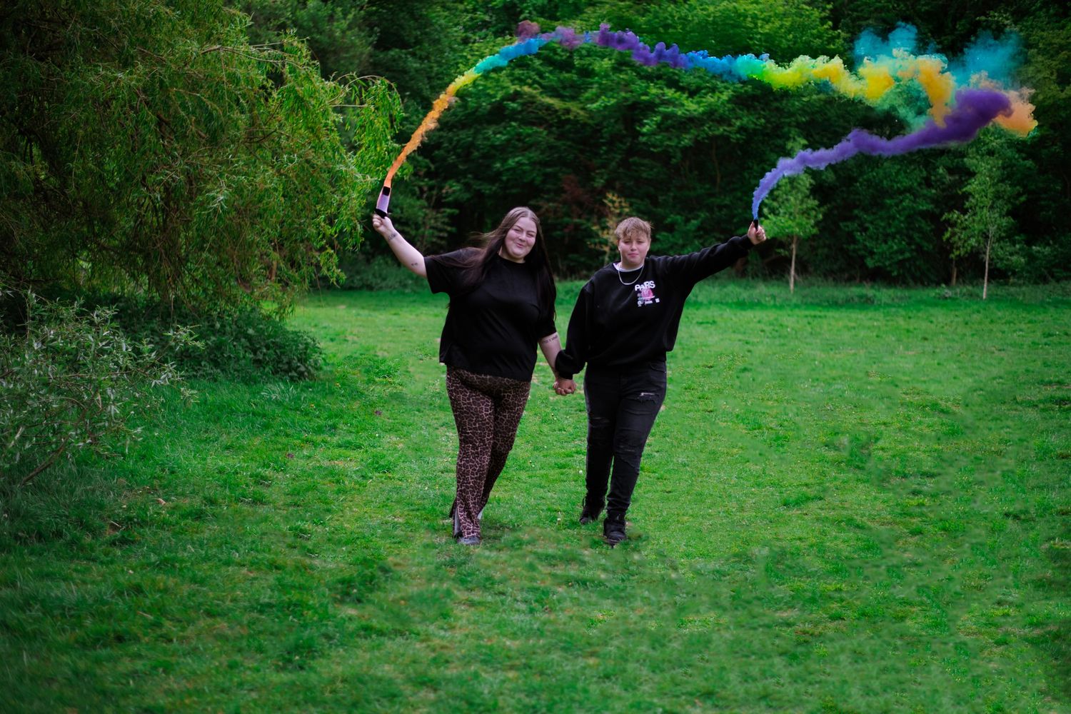 Two people run through a green field holding colorful smoke flares that create rainbow trails behind them in a vibrant display.