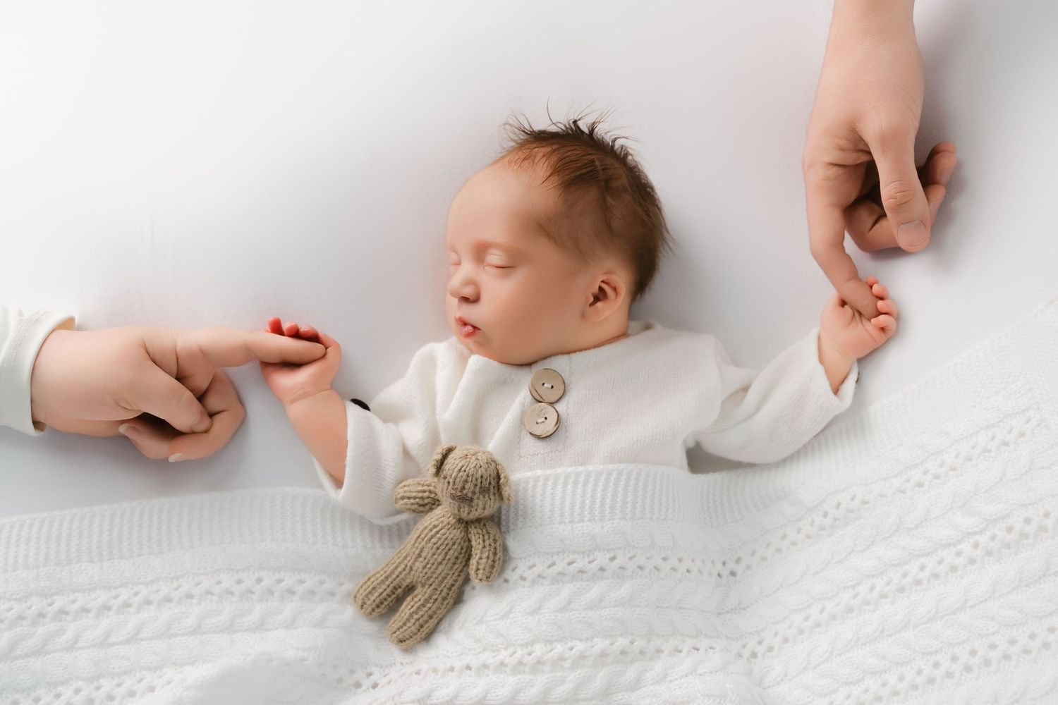 A sleeping baby in white clothing holds hands with someone while clutching a small teddy bear on a textured white blanket.