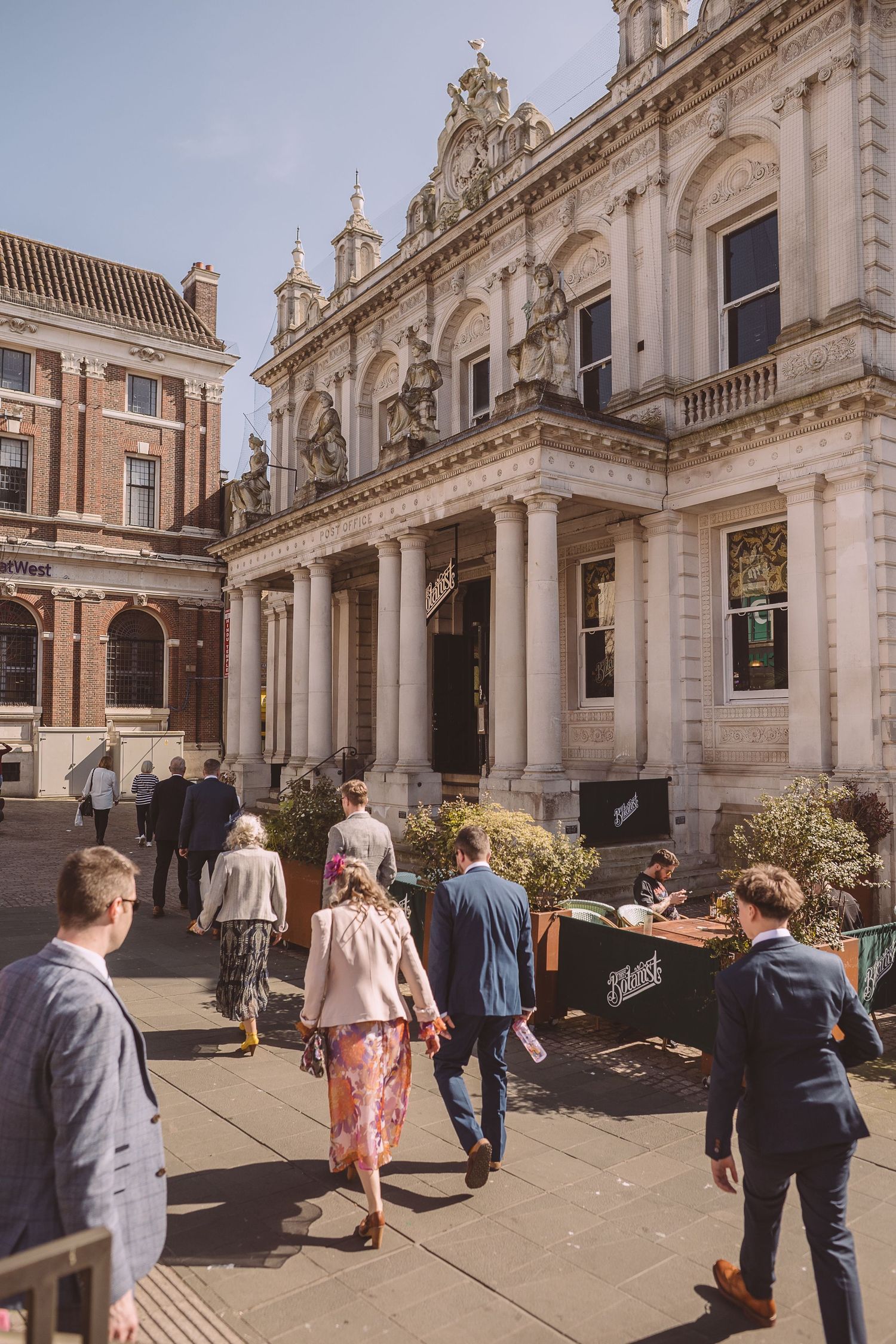 People gathered outside a grand classical-style building with ornate stone columns and architectural details in warm sunlight.