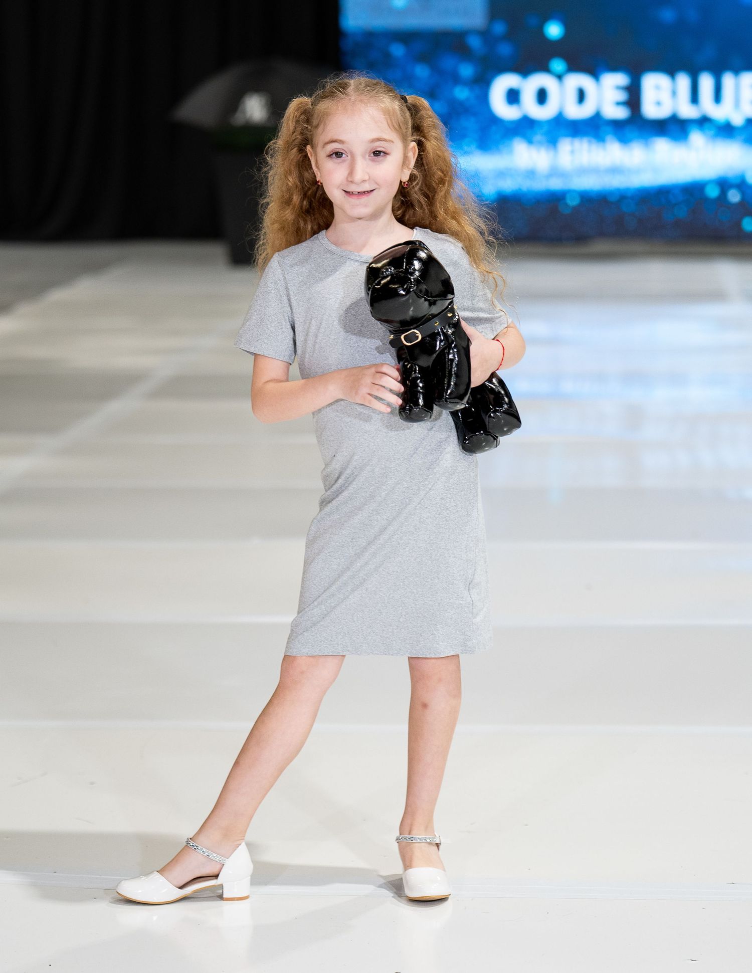Young performer in a gray dress holds a black plush toy while walking a runway during a fashion show event.