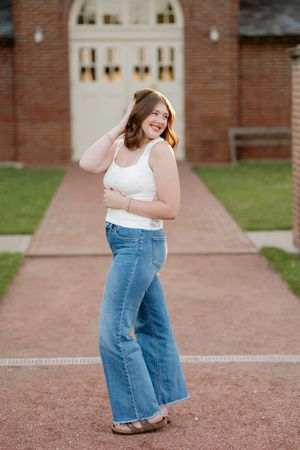 Young woman in white tank top and wide-leg jeans posing outdoors on brick pathway near historic building.