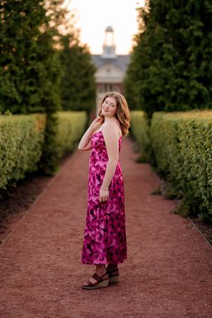 Young woman in pink floral maxi dress poses on garden path flanked by tall hedges at golden hour.