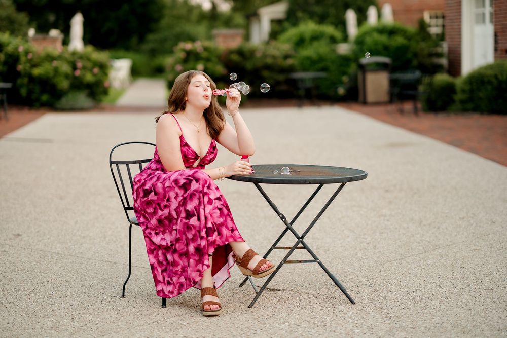 Young woman in pink floral maxi dress blowing bubbles at outdoor cafe table in garden setting.