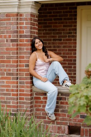 Young woman in pink top and light jeans sits smiling on brick porch ledge wearing white sneakers.