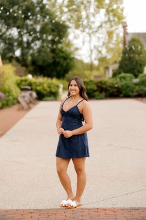 Young woman in navy blue mini dress and white sandals smiling outdoors on brick path surrounded by greenery.