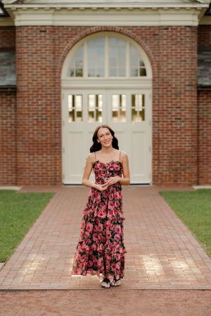 Woman in floral maxi dress standing on brick path before arched door of elegant brick building.