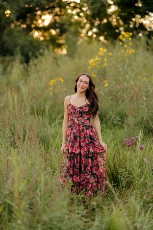 Young woman in floral red maxi dress standing in a lush green meadow with wildflowers at golden hour.