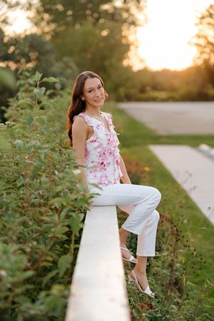 Young woman in floral top and white pants sits on a garden wall at golden hour sunset.