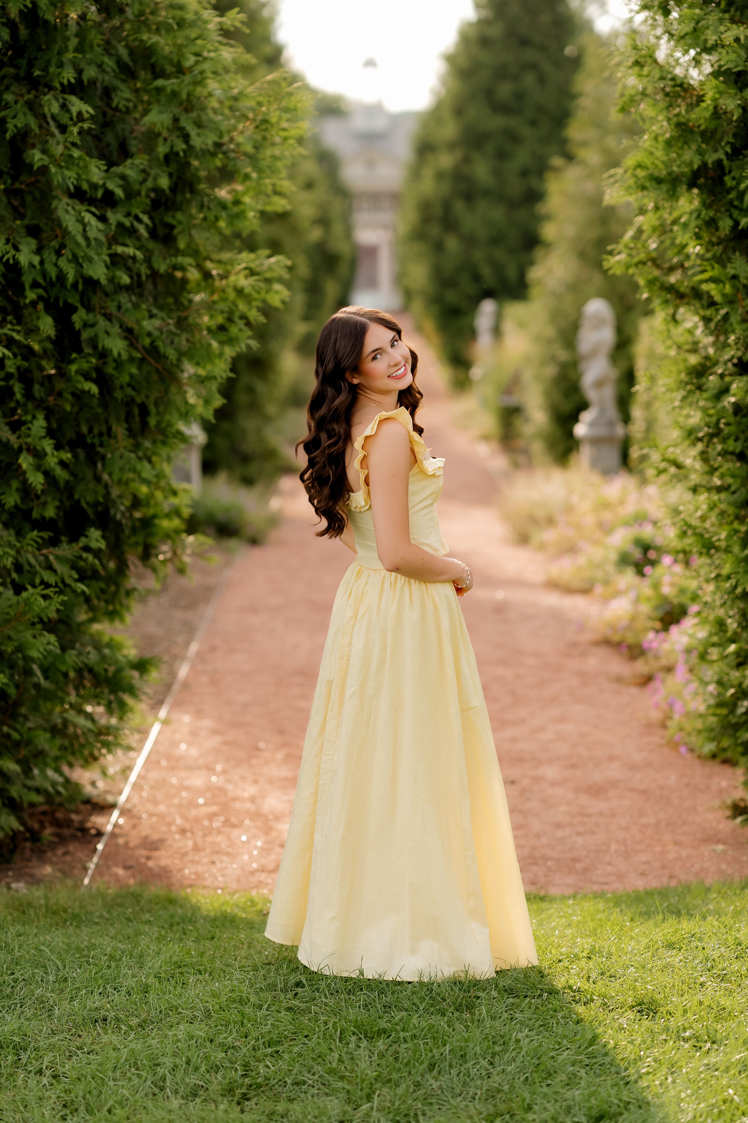 Young woman in elegant yellow ball gown smiling over shoulder in lush garden pathway with tall hedges.