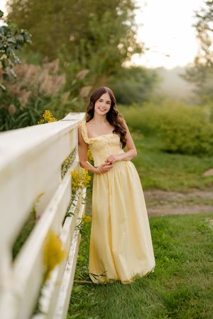 Young woman in yellow maxi dress leaning on white fence surrounded by lush greenery during golden hour.
