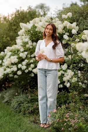 Young woman in white blouse and jeans holding flowers beside blooming white hydrangea bushes in a garden.