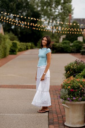 Young woman in mint top and white maxi skirt smiling outdoors with string lights and flower planters.
