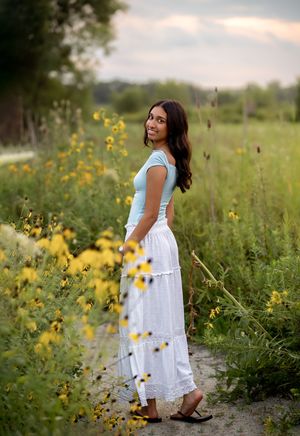 Young woman in light blue top and white maxi skirt standing on a path surrounded by yellow wildflowers.