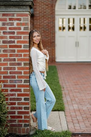 Young woman in white crop top and light blue jeans leaning against a brick pillar, smiling outdoors.