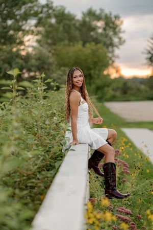 Young woman in white dress and cowboy boots sitting on a fence surrounded by wildflowers at sunset.
