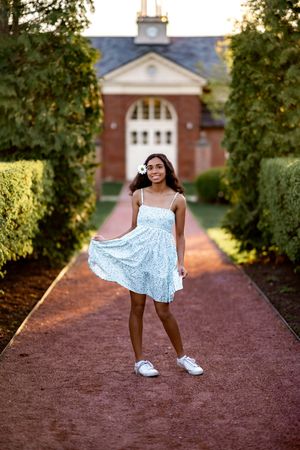 A person in a light blue summer dress poses on a brick pathway leading to a stately brick building with white columns.