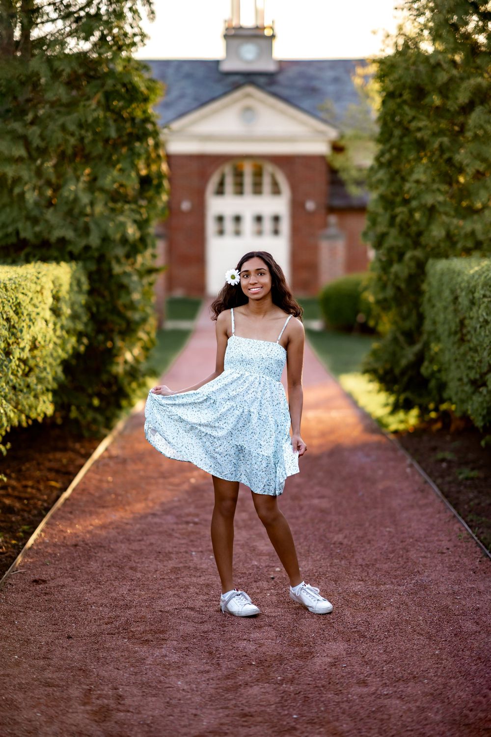 A person in a light blue summer dress poses on a brick pathway leading to a stately brick building with white columns.