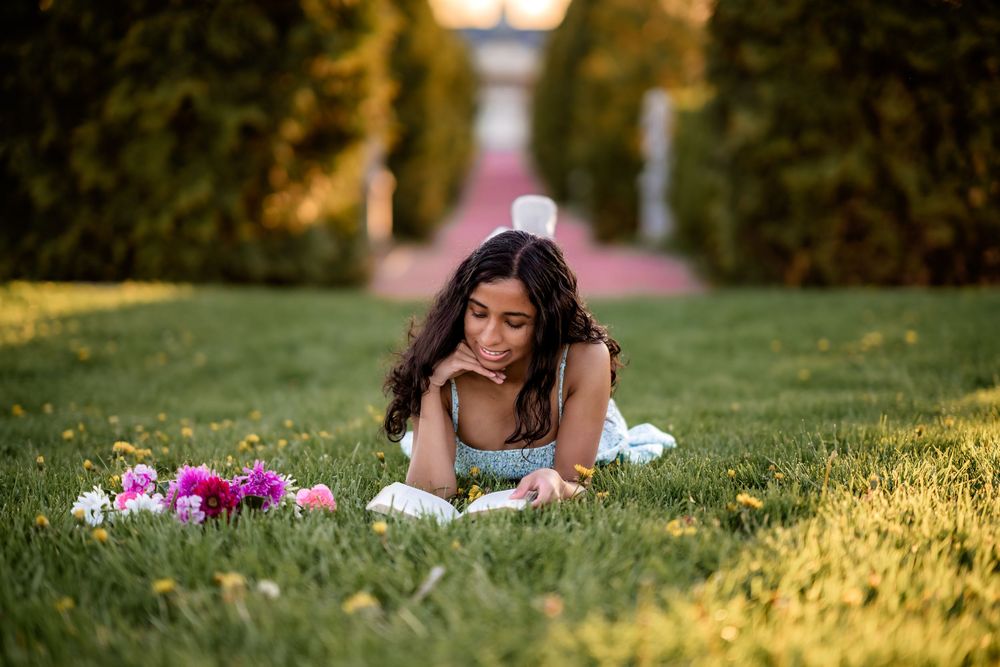 A person lies on grass reading a book beside pink flowers in a peaceful garden setting at golden hour.