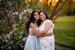 Two people share an embrace while standing next to flowering bushes during a golden sunset in a park.