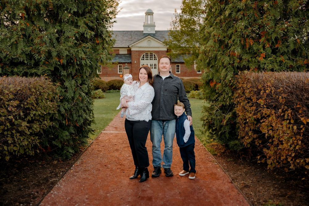 A family stands together on a brick path surrounded by autumn trees and fallen leaves.