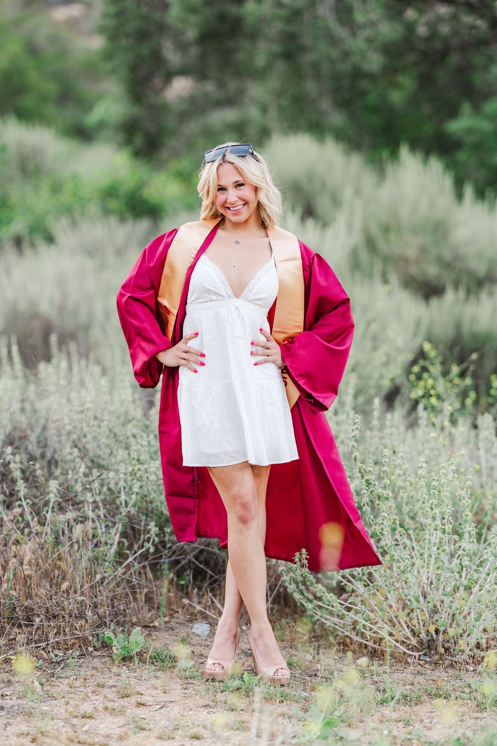 Graduate wearing a white dress and burgundy graduation gown poses outdoors in a natural desert landscape setting. Graduation Photos