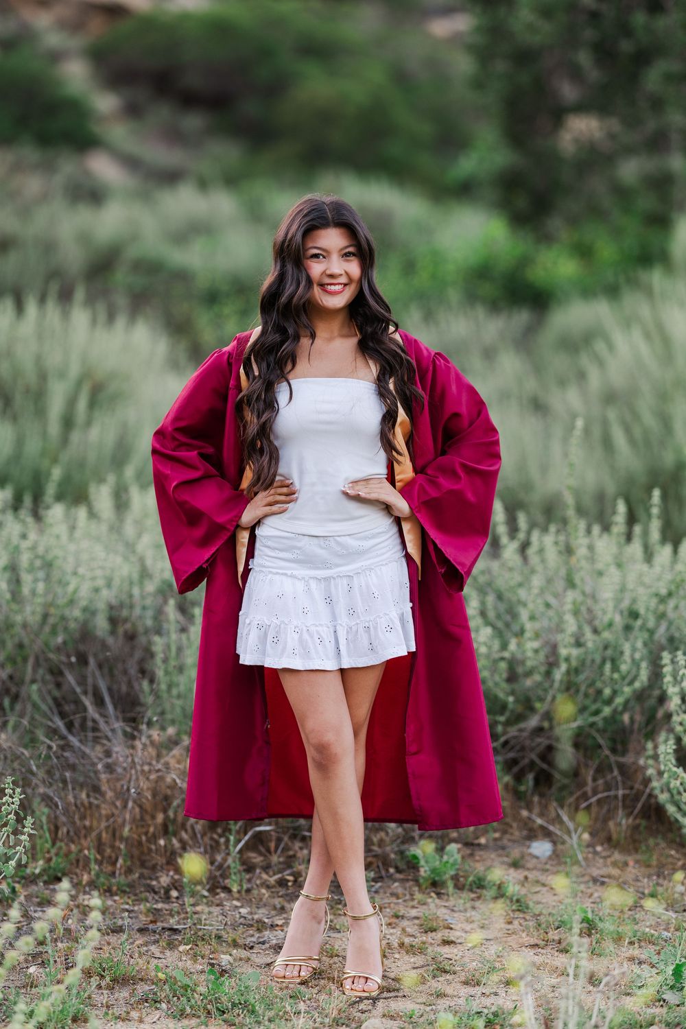 A figure in a white dress and burgundy coat poses outdoors against green foliage. Graduation Photos
