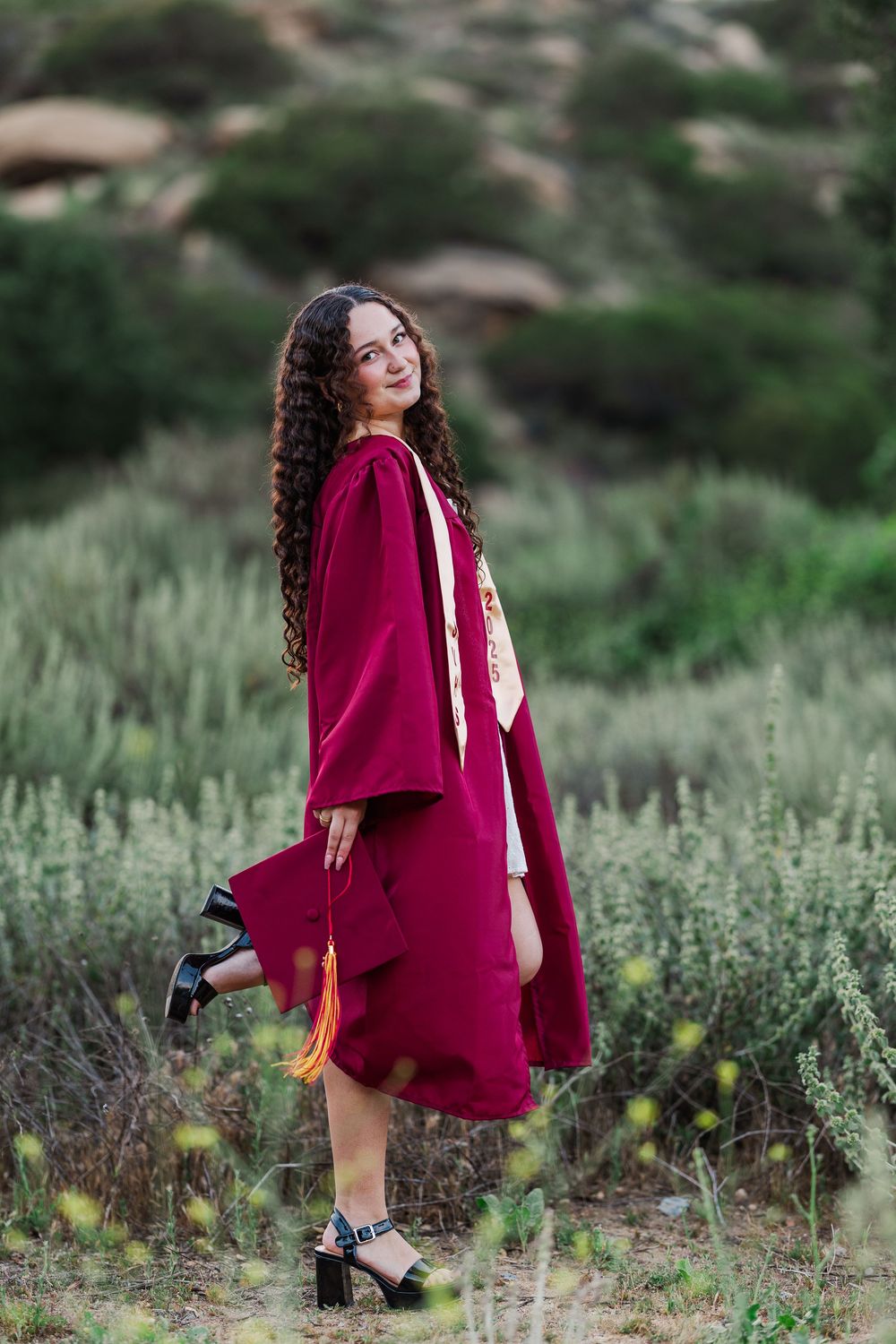 A person in a burgundy graduation gown stands in a meadow with long curly hair and block heels. Graduation photos