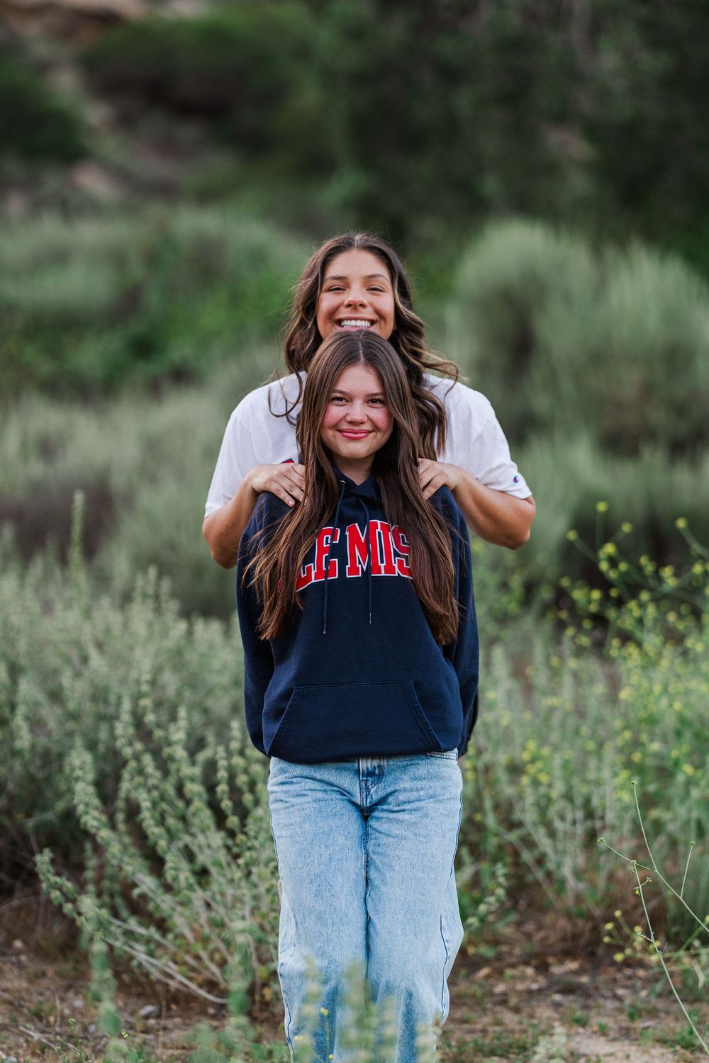 Two friends smiling and laughing together outdoors among green foliage while wearing casual clothing.