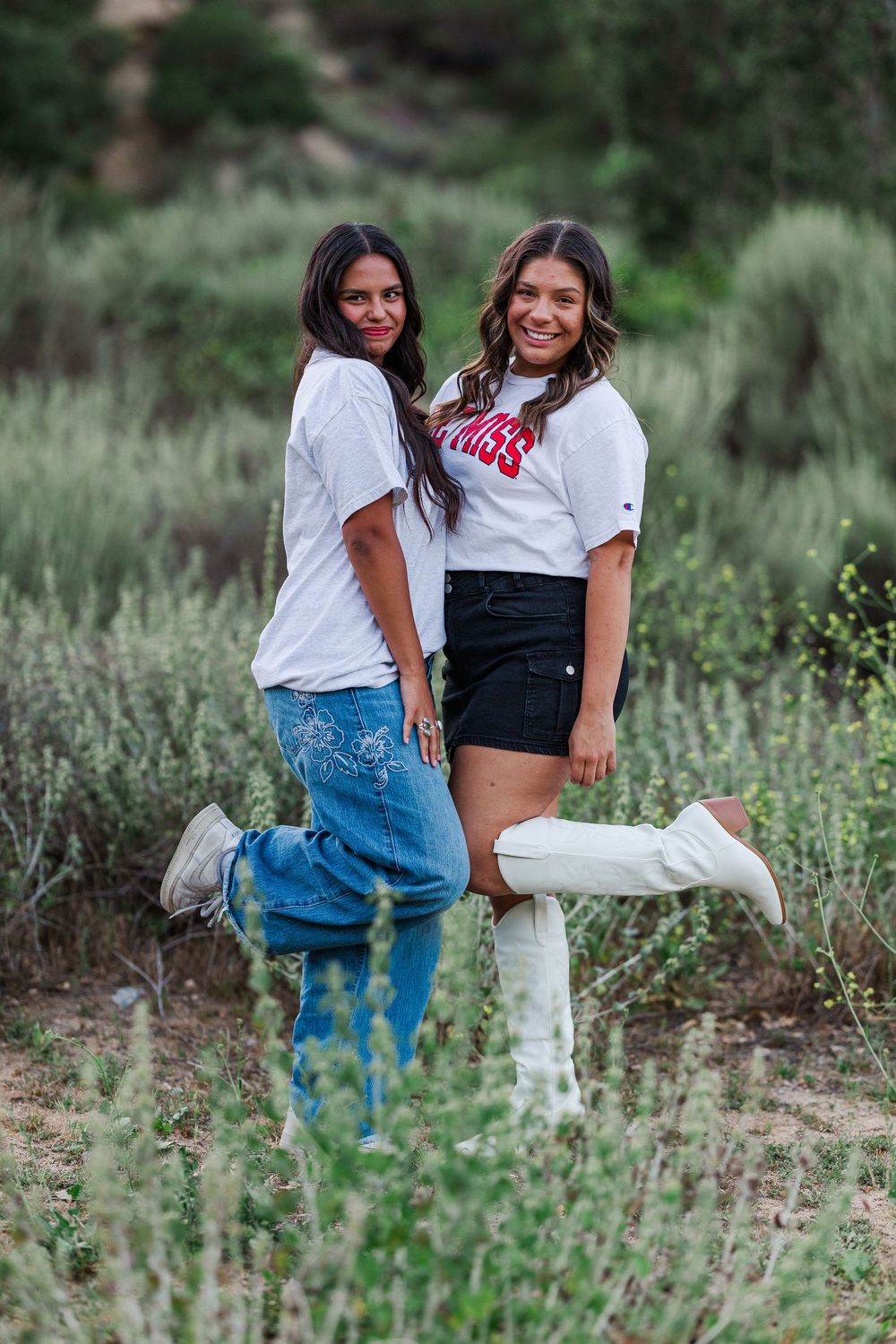 Two friends pose in casual outfits and white sneakers among tall grasses in an outdoor nature setting.